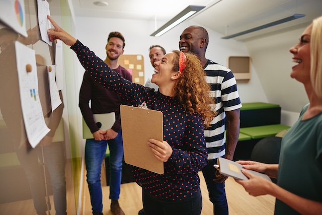 Smiling group of diverse work colleagues reading charts and graphs taped to a wall during an office meeting for Turning Point Licensed Behavior Analyst Services, PLLC